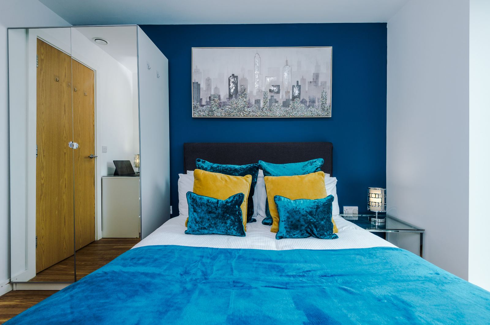 Styled bedroom with deep blue accent wall, velvet cushions and city skyline canvas
